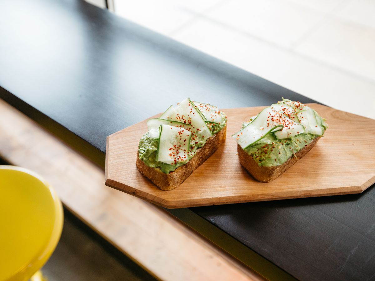 Two slices of avocado toast with cucumber and a sprinkle of red spice sit on a wooden board in a window. 