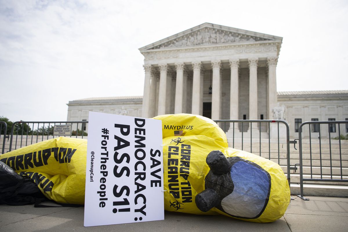 An inflatable figure wearing a yellow hazmat suit and gas mask lies on its side carrying a sign that reads “Save democracy, Pass S 1.” The Supreme Court building is in the background.
