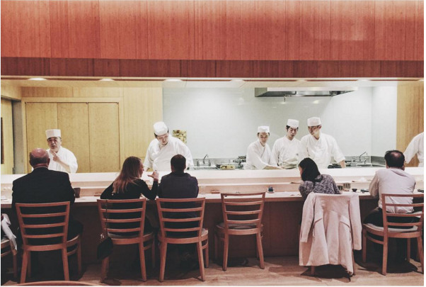 A handful of guests sit at a sushi counter, while multiple people in a white chef’s outfit and hat work behind the counter