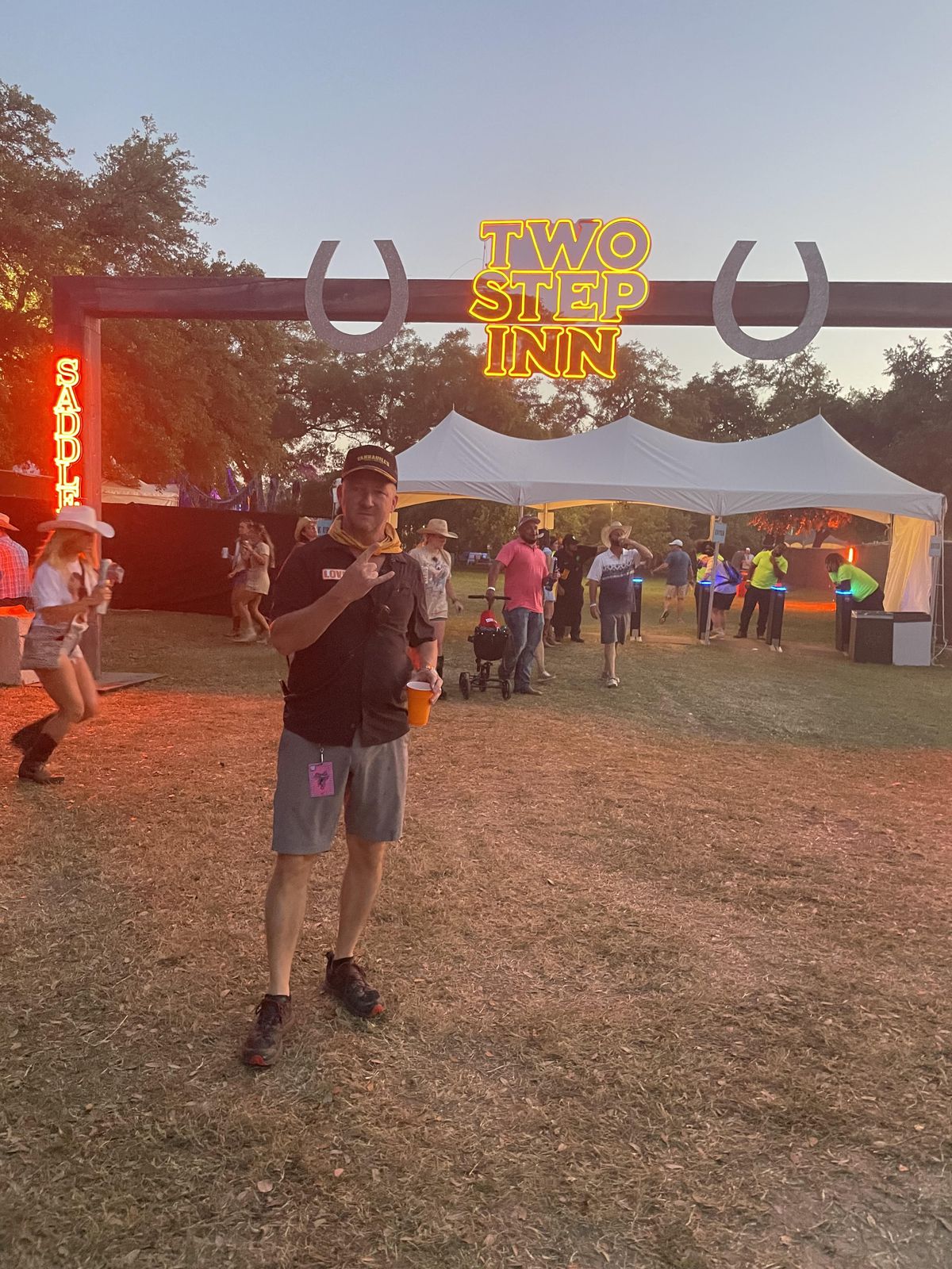 Tim Love stands in front of the entrance to the Two Step Inn music festival, with a neon sign lit up behind him and many people on the festival grounds.