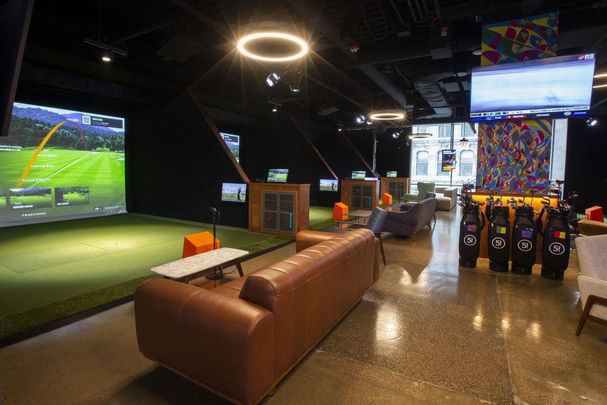 A row of large golf simulator bays with putting greens and large screens in font of couches.
