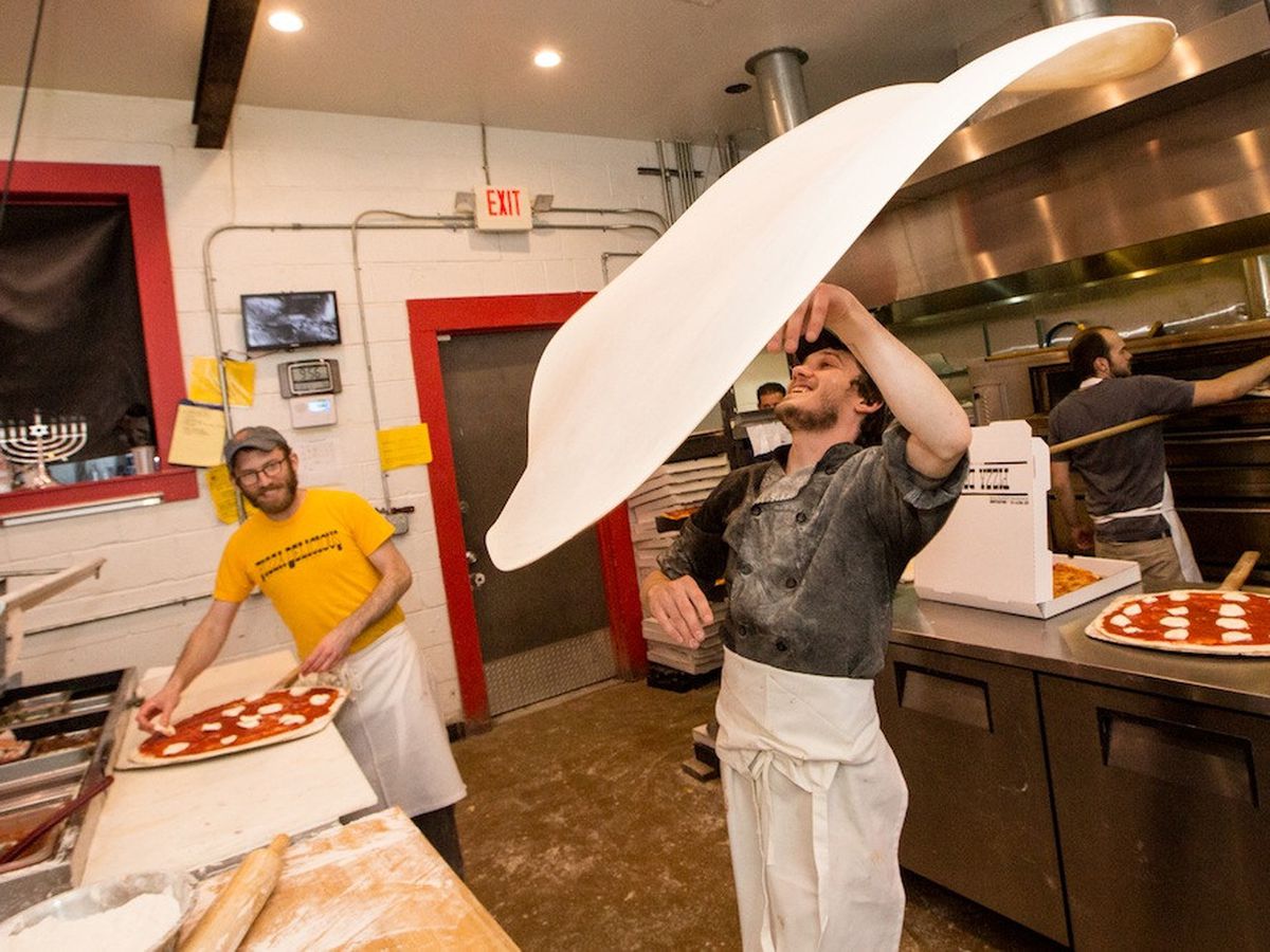 A person wearing a grey shirt and a white apron throwing a large round of pizza dough in a kitchen, with another person smiling in the background.