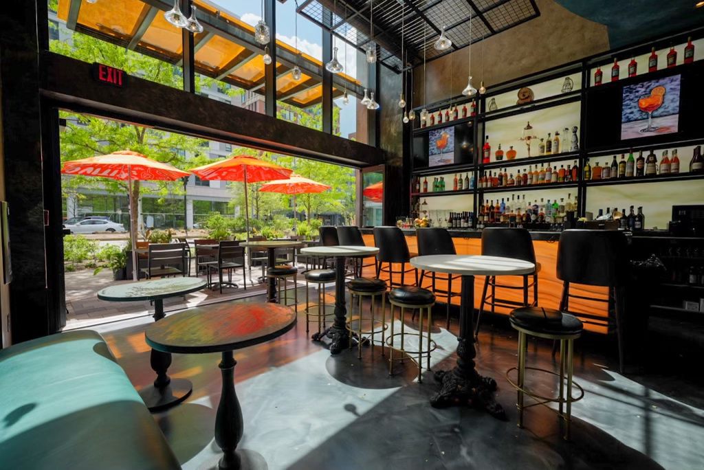 A bar with the front windows rolled up to show an outdoor patio with orange umbrellas shielding tables from the sun.
