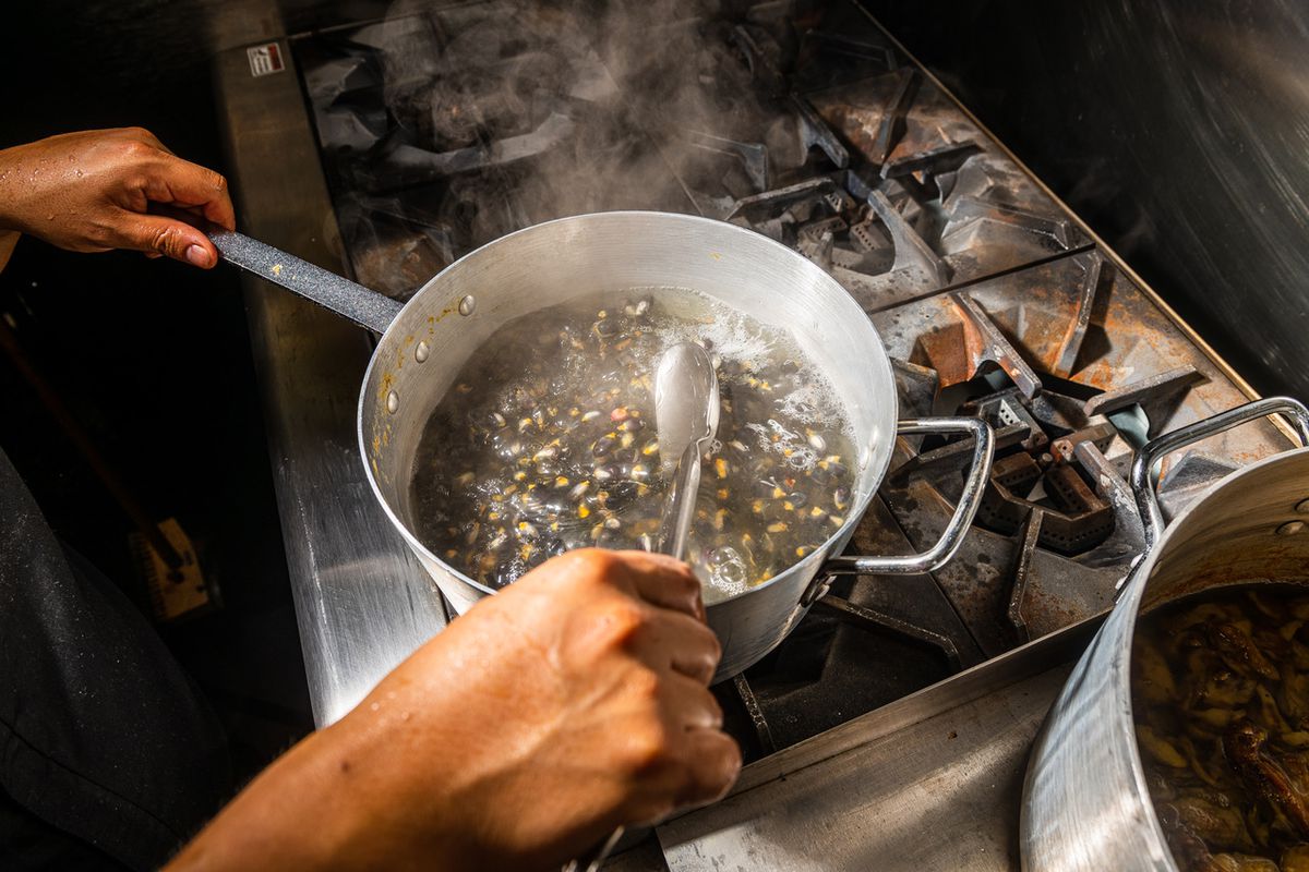 A pair of hands stirs a pot of blue and yellow corn simmering on a stove at Maïz64.