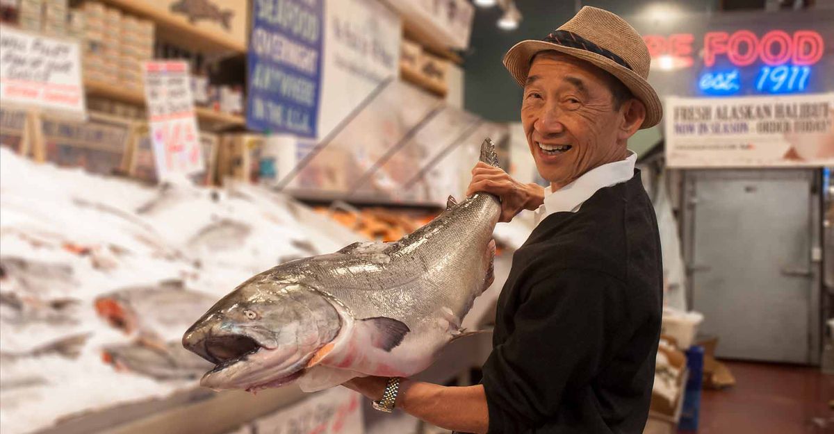 A man wearing a straw fedora holds an entire salmon at a fish market while smiling.