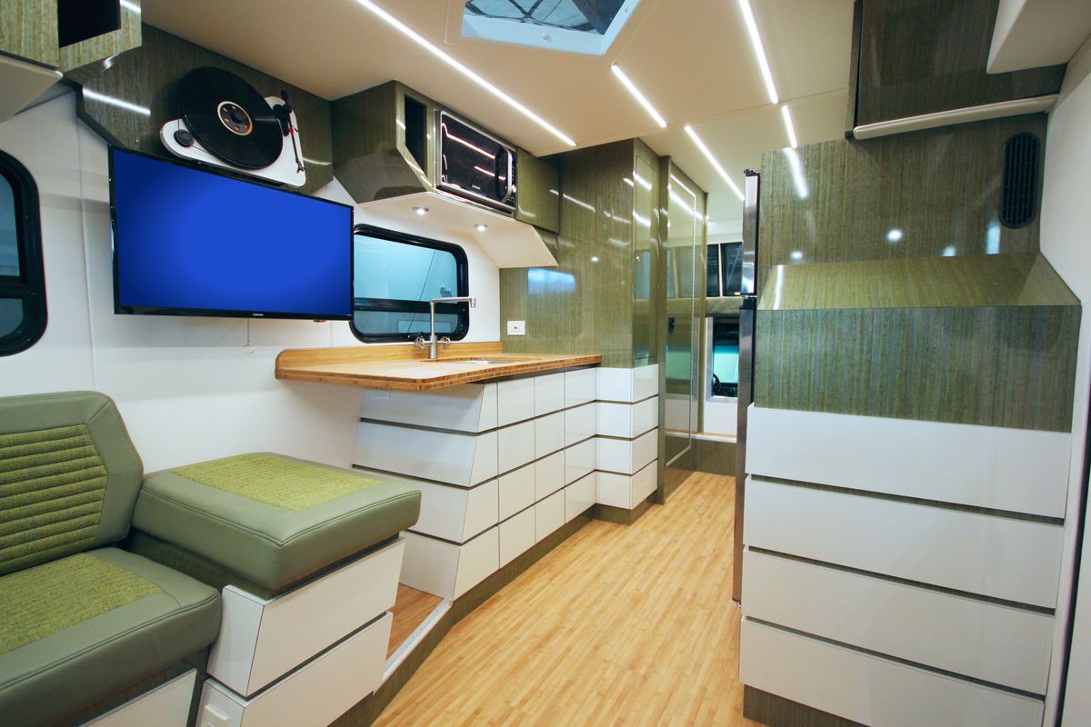The interior of a camper has white cabinets, green accents, wood floors, bamboo countertops, a TV, and a hallway towards the front cab. 