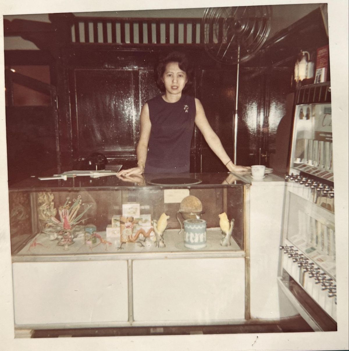 An old photograph of a woman wearing a blue dress, standing behind a glass counter.