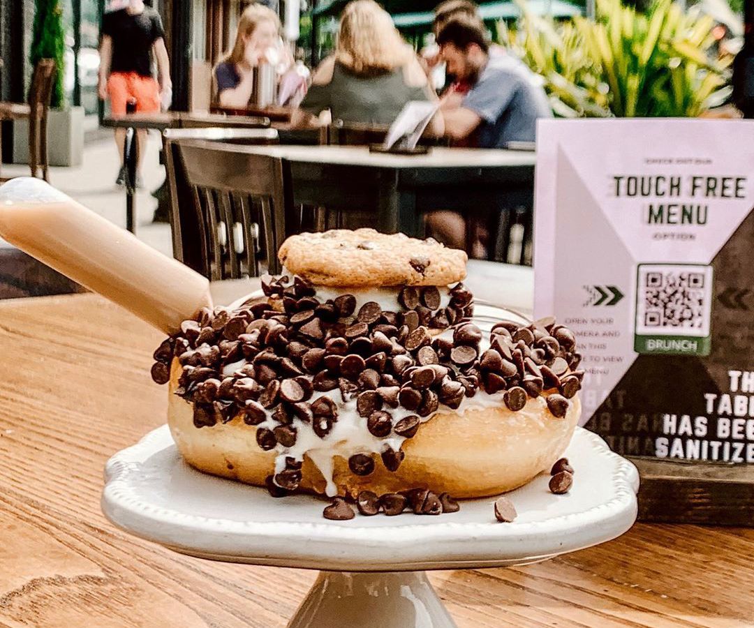 A doughnut / chocolate chip cookie sandwich oozing with chocolate chips and cream sits on a patio table. A flyer behind it advertises a touch-free ordering process.