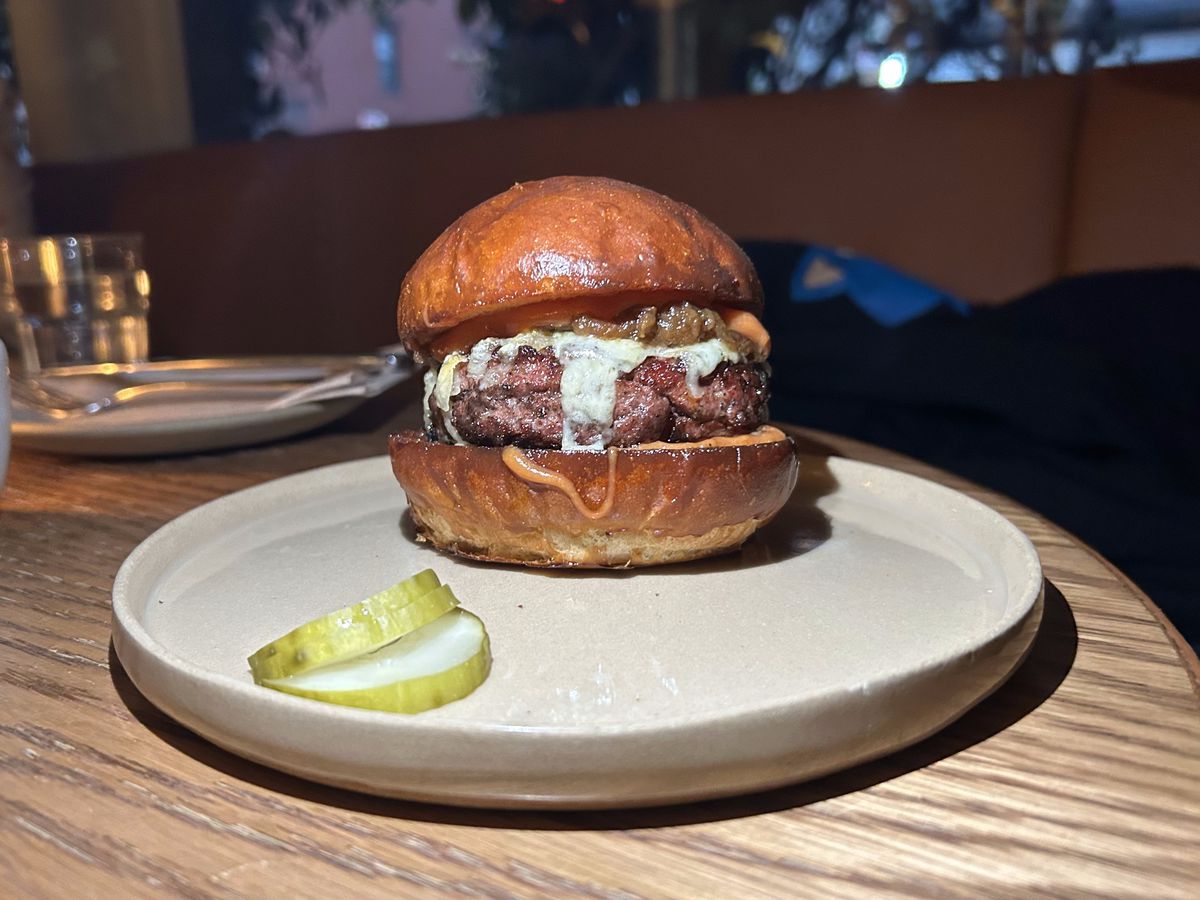 A burger dripping with white cheese and caramelized onions sits on a plate next to three half-slices of pickle.