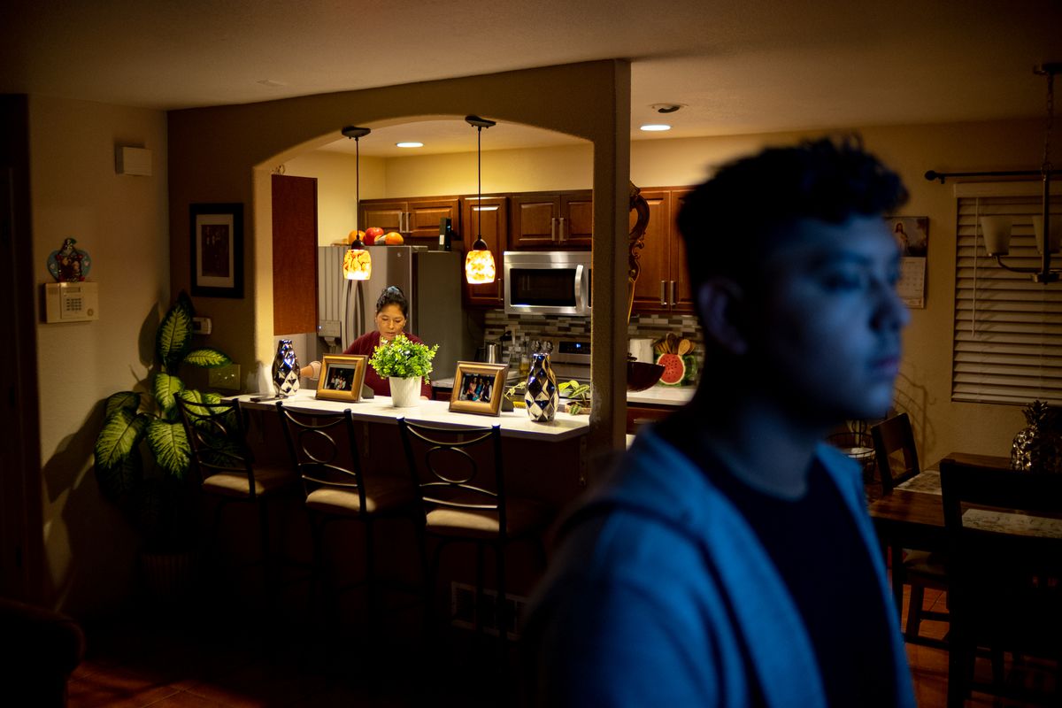 A mother works in the kitchen during an early morning as her son looks at the television in the living room, its blue light reflecting off of his face.