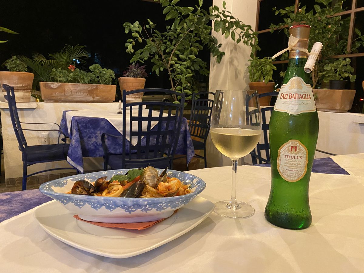 A decorative dish of pasta topped with mussels and other seafood, beside a bottle of wine on a white tablecloth lined table