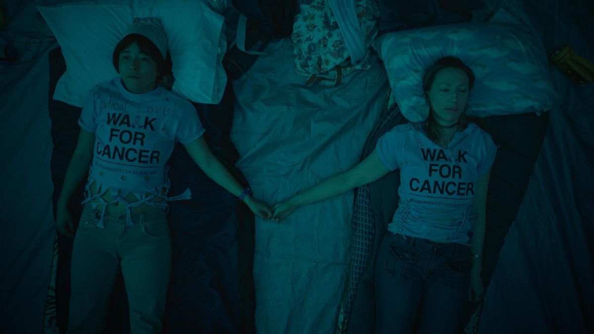 Maya Erskine and Anna Konkle playing tween versions of themselves in Pen15. The two are wearing Walk for Cancer shirts, and holding hands, while laying on top of sleeping bags, in a tent.