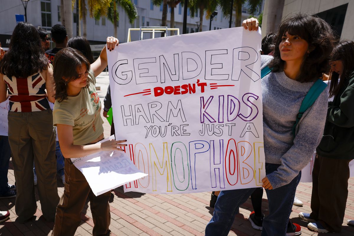 Two high school students hold a sign reading, “Gender doesn’t harm kids, you’re just a homophobe.”