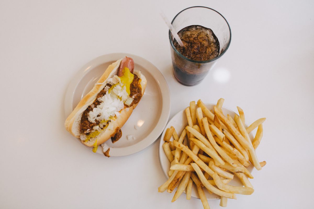 A coney dog with mustard, chili, and chopped white onions next to a plate of plain french fries and a cup of brown soda with ice and a straw.