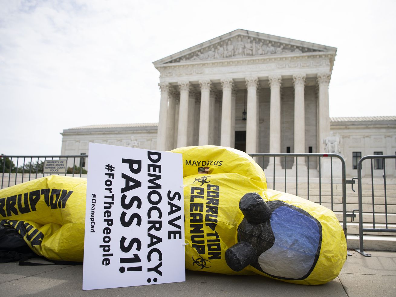 An inflatable figure wearing a yellow hazmat suit and gas mask lies on its side carrying a sign that reads “Save democracy, Pass S 1.” The Supreme Court building is in the background.