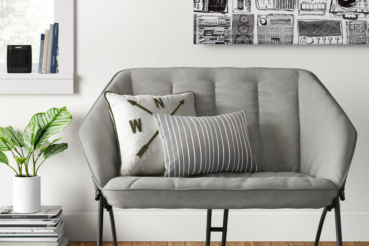 A small gray foldable sofa with two pillows sits next to a stack of magazines and a houseplant.