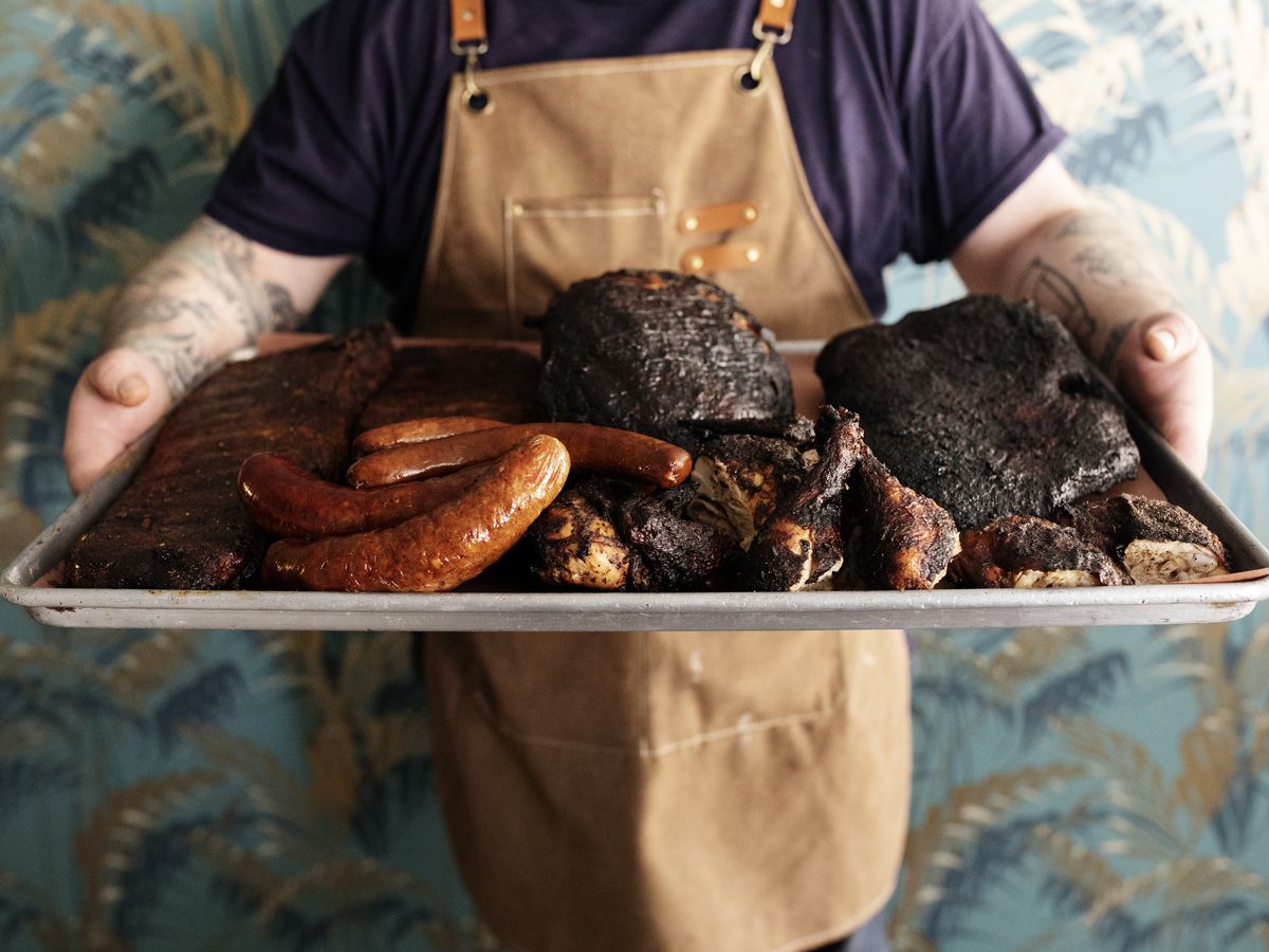 A person wearing an apron holding a tray of various smoked meats. 