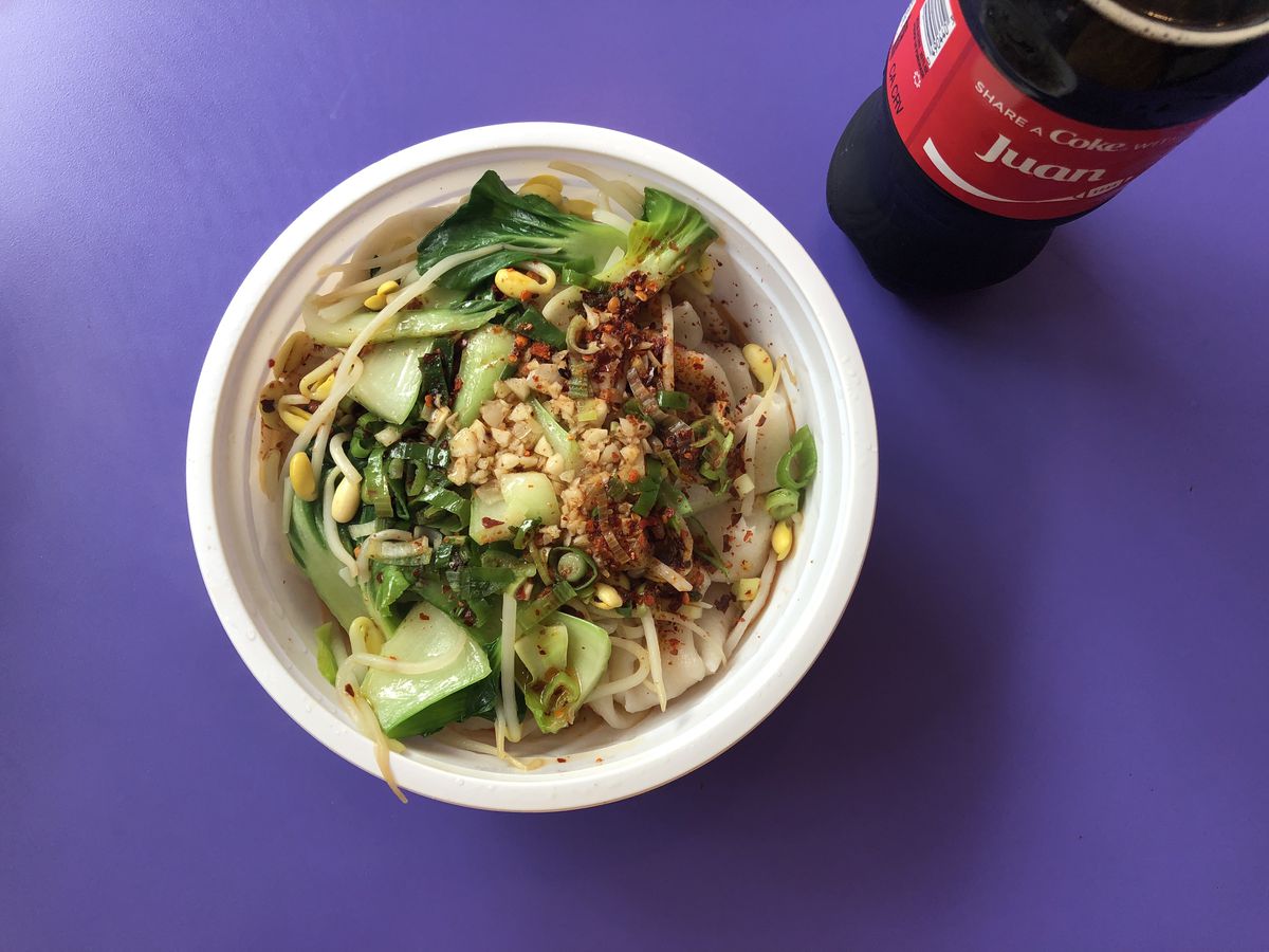 A white plastic bowl, sitting on a purple table, is filled with hand-pulled noodles, bok choy, chile flakes, and bean sprouts. A plastic Coke bottle appears to the side.