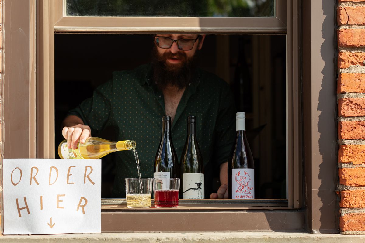 A man with a beard and glasses pouring wine into a glass from inside a window.