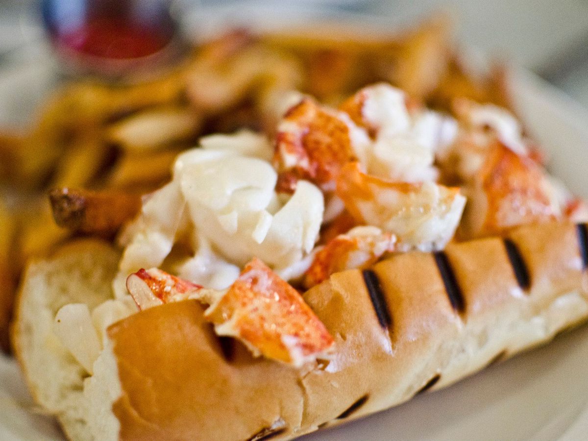 The Maine lobster roll at Neptune Oyster is served on a grilled hot dog bun atop a white plate, and is accompanied by French fries and a ramekin of ketchup.