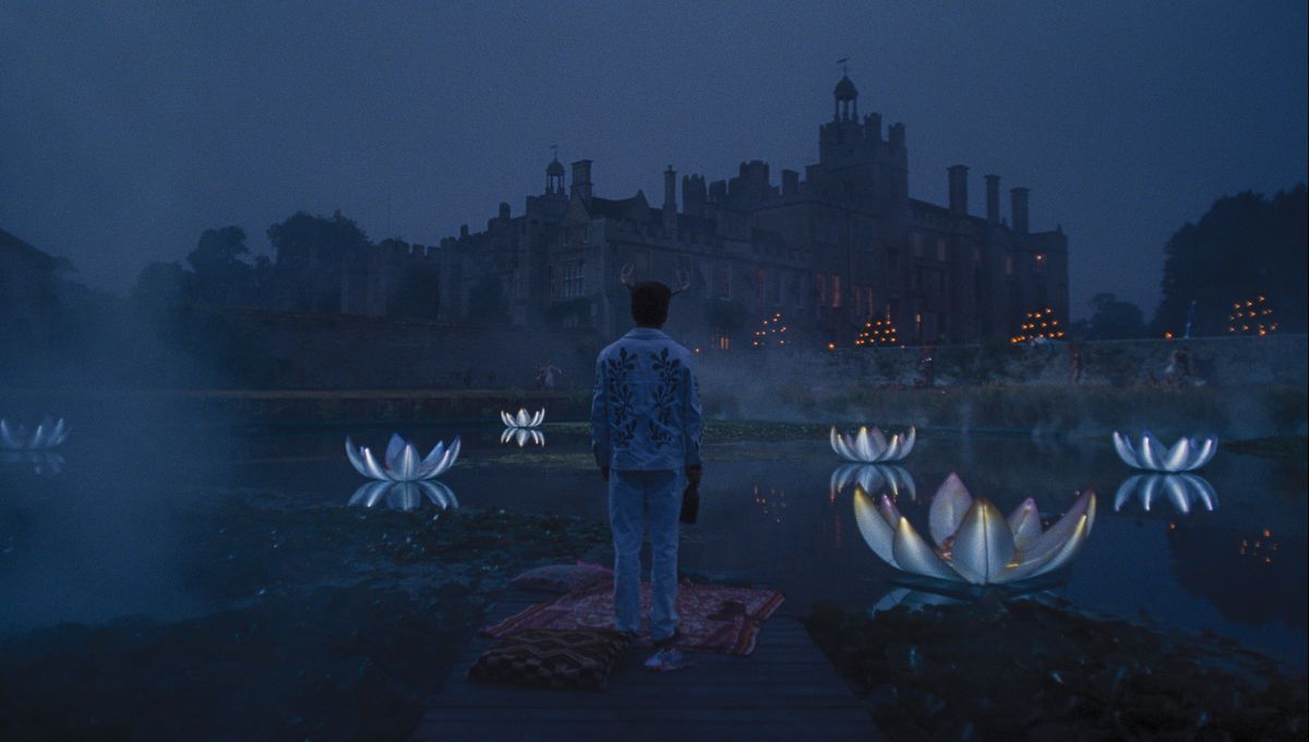 Oliver (Barry Keoghan), dressed for a costume party in deer horns and an elaborately beaded white suit, stands on a blanket on a dock with his back to the camera, looking out over a pond full of huge light-up floating plastic lilies, and beyond them, an immense Gothic estate, in the movie Saltburn