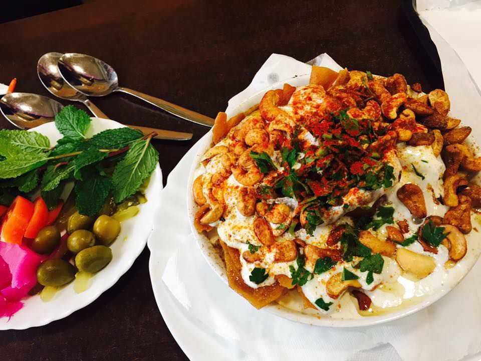 A bowl of fatteh topped with lots of fixings, including herbs, nuts, powdered spices, and pita chips