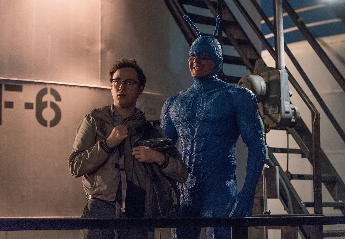 The Tick grins widely as Arthur looks confused, while standing in front of a stair case and a silo in The Tick.