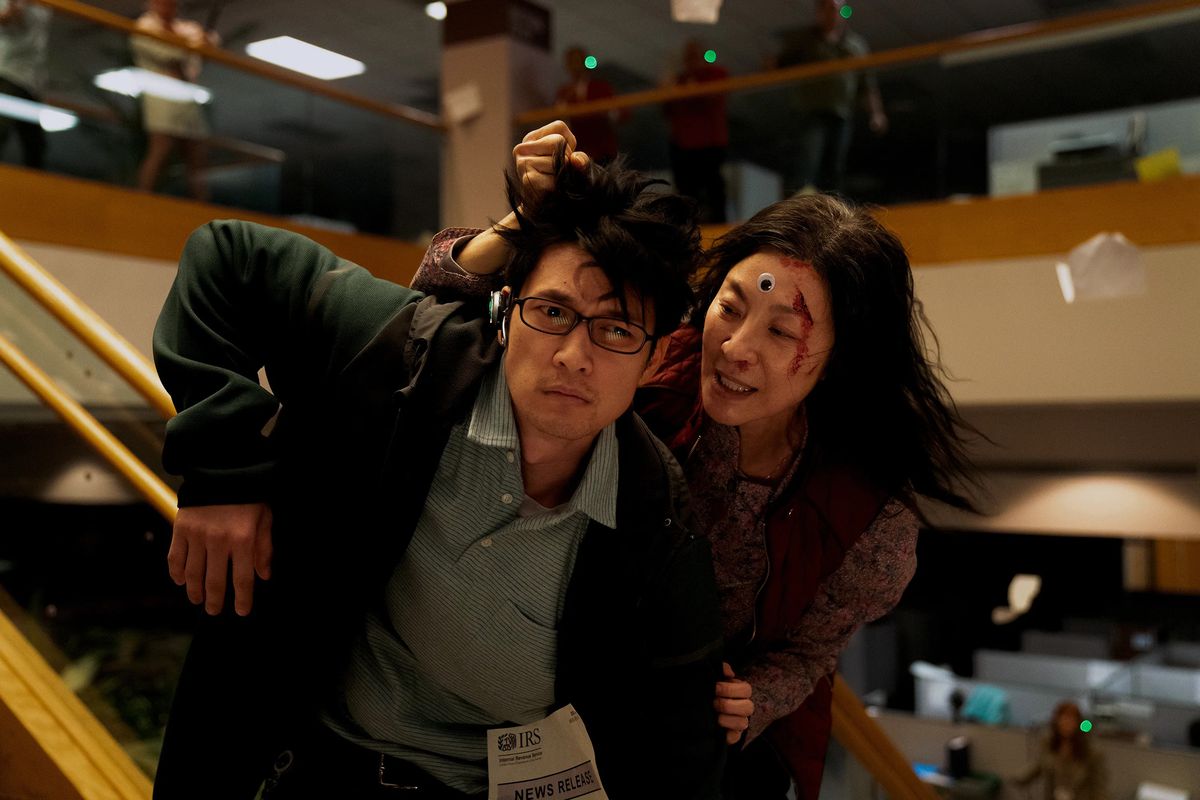 A woman with a googly eye stuck to her forehead holds a handsome younger man in glasses by the hair and seems to be speaking into his ear. They both seem to have recently been through a tussle, and they’re in an office building.