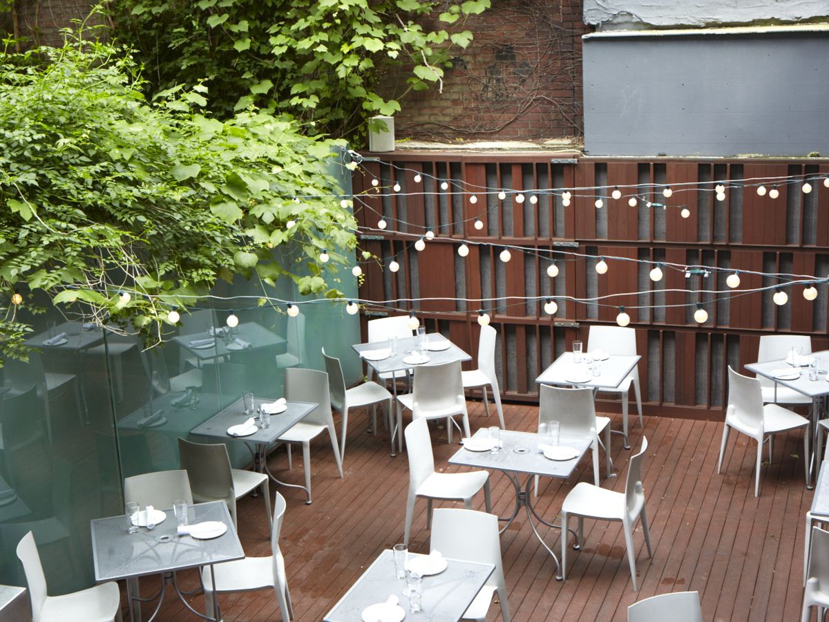 Light-colored tables and chairs sit on a nice wooden restaurant patio with trees coming over one wall and string lights above.
