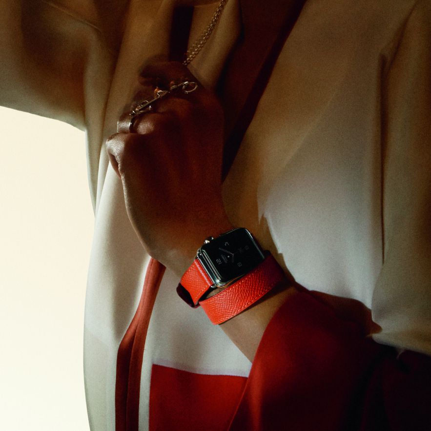 A female model dressed very retro-seventies wears an Apple Watch with an orange Hermès band. 