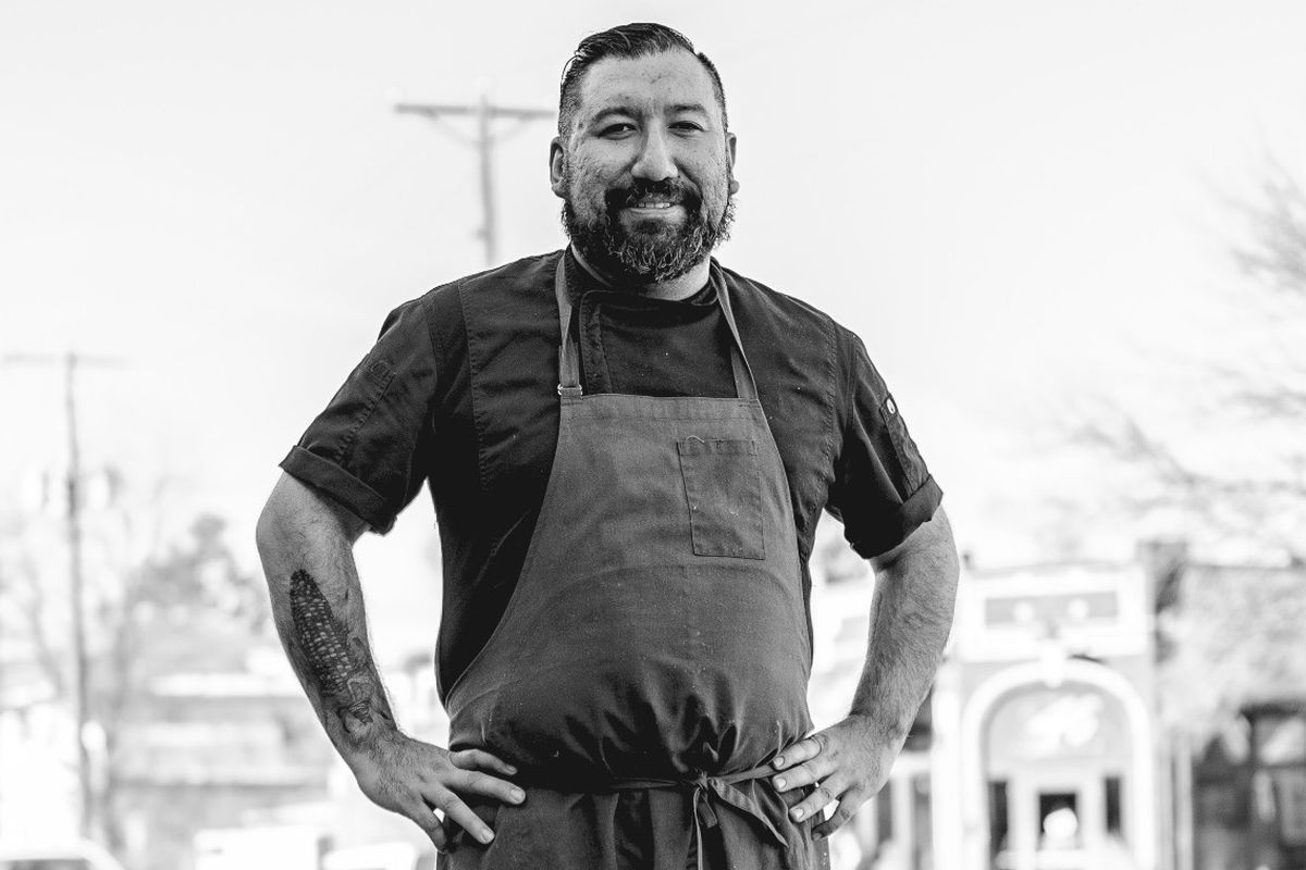 Black and white image of Gustavo Romero from the torso up, hands on his hips, wearing a chef coat and an apron. A corn cob tattoo is visible on his forearm.
