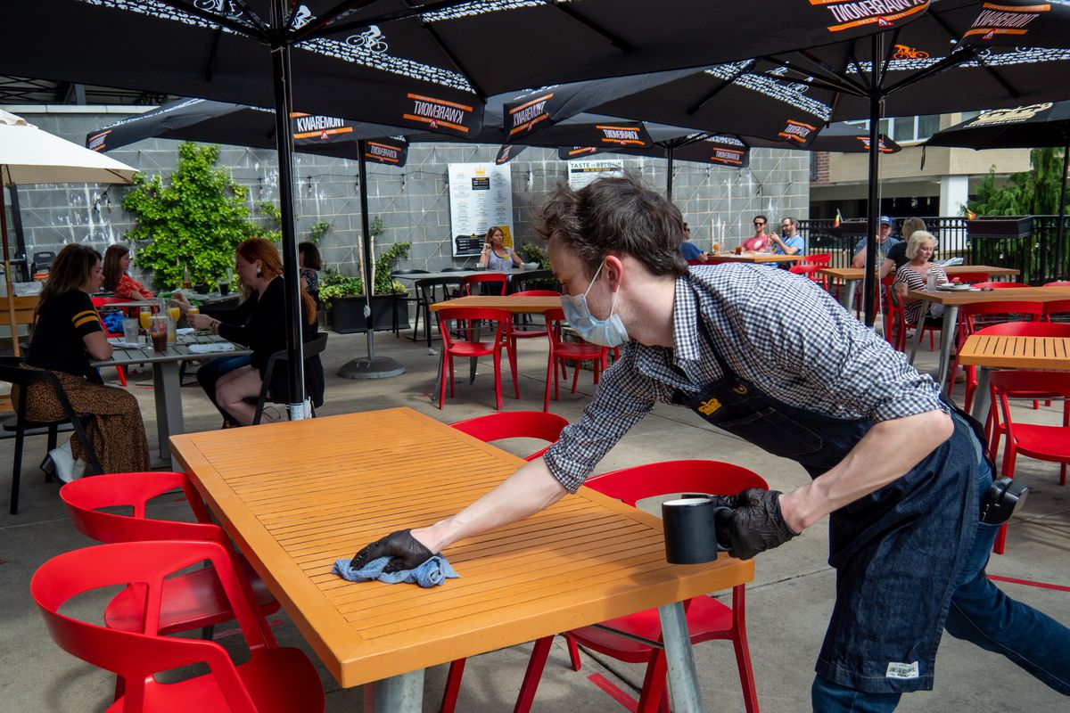 A worker in gloves and a mask wipes down an outdoor table with red chairs.