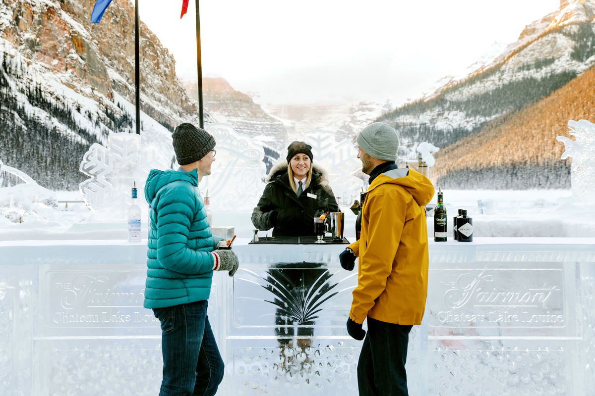 Two customers stand a bar made of ice with a bartender. Flags fly behind, and there are sunlit mountains beyond