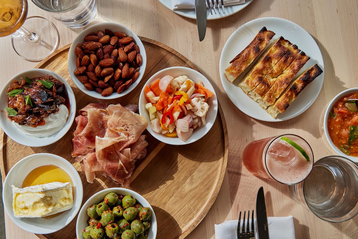 A collection of small plates on a wooden tray, with a side of focaccia and assorted drinks.