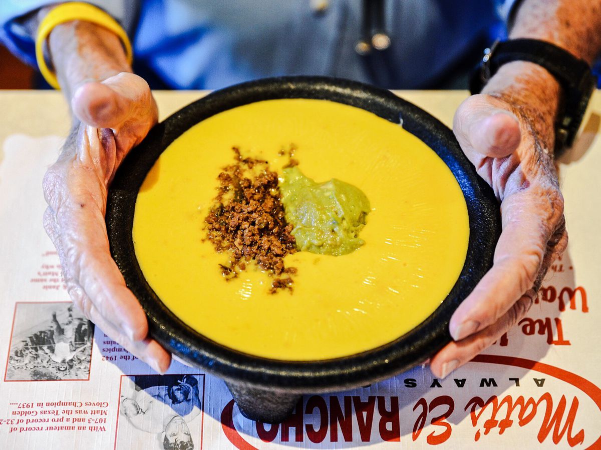 A person holding up a big bowl of yellow melted cheese with a clump of ground meat and guacamole in the middle.