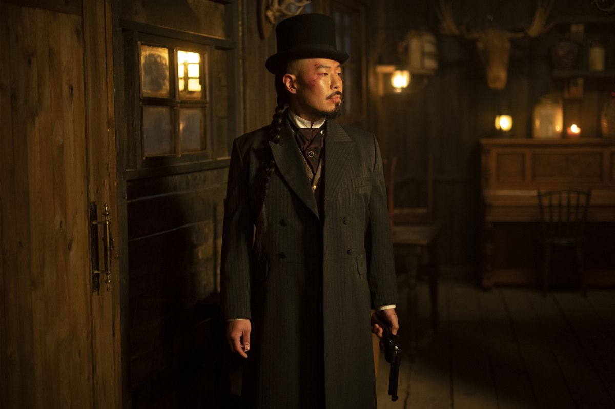 Hoon Lee, wearing a suit with a top hat, stands in front of a closed door in Warrior.