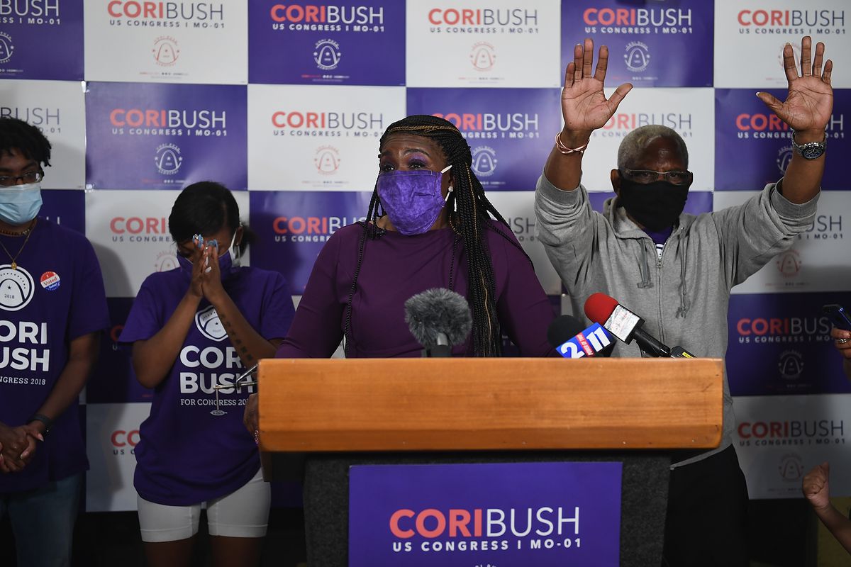 Congressional candidate Cori Bush wears a mask while giving a speech from behind a podium. Campaign supporters stand behind and to the sides.