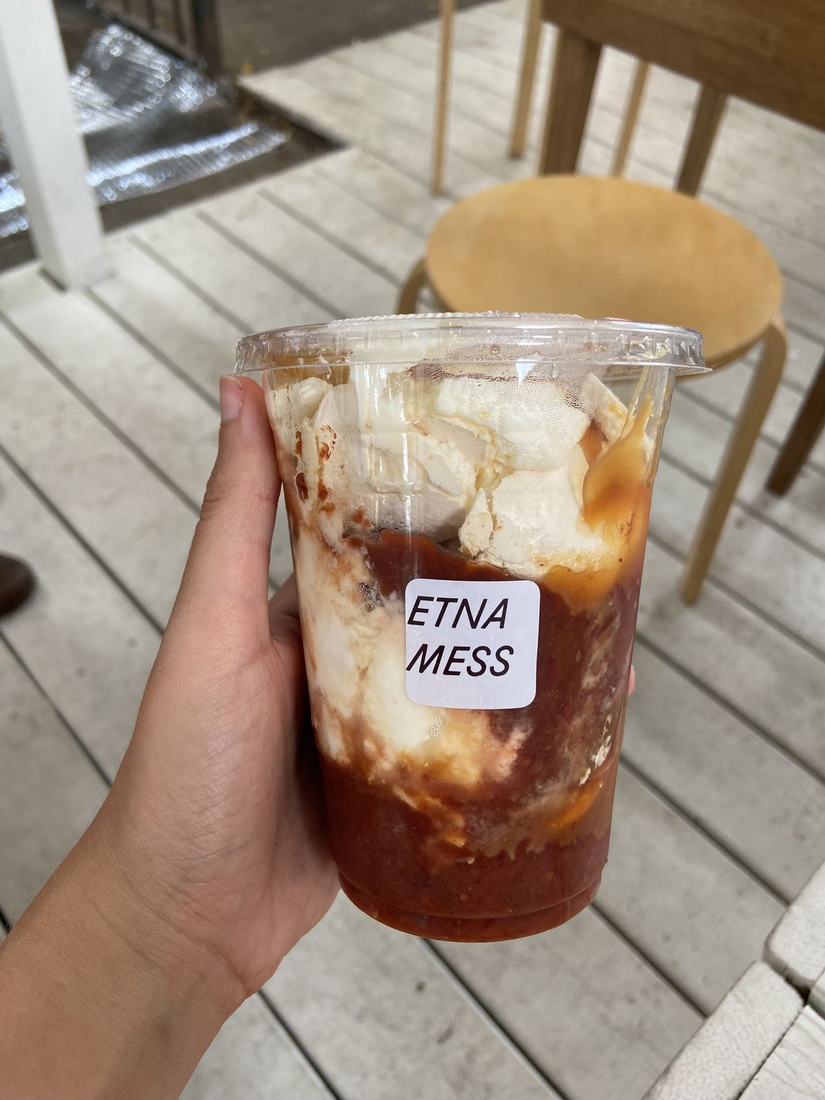 Hand holding a plastic cup labeled Etna Mess filled with tomatoes, strawberries, meringues and caramel.