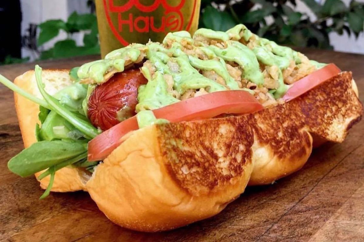 a close-up of a hot dog covered in smashed avocado, with a pale beer in a frosty glass behind it