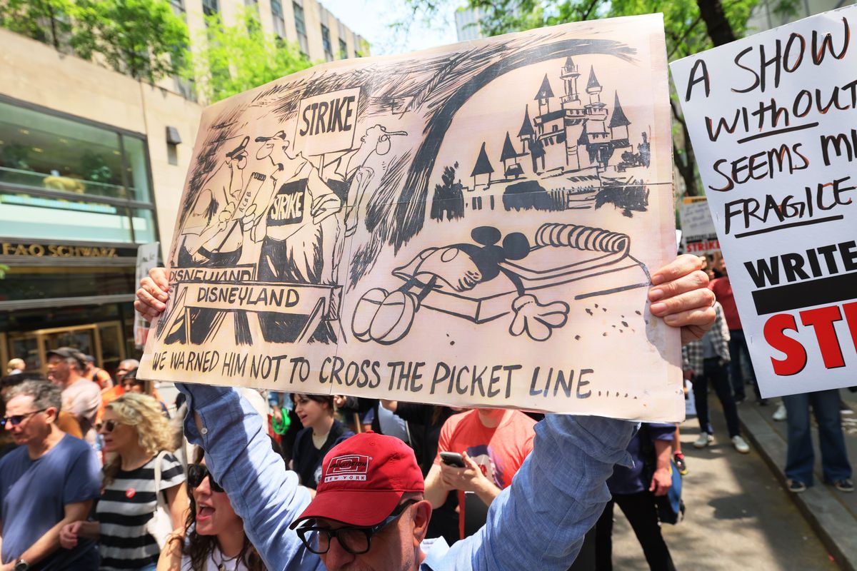 A protester holds a sign with a cartoon of people speaking on one side and Mickey Mouse caught in a spring trap on the other. The caption reads, “I warned him not to cross the picket line.”