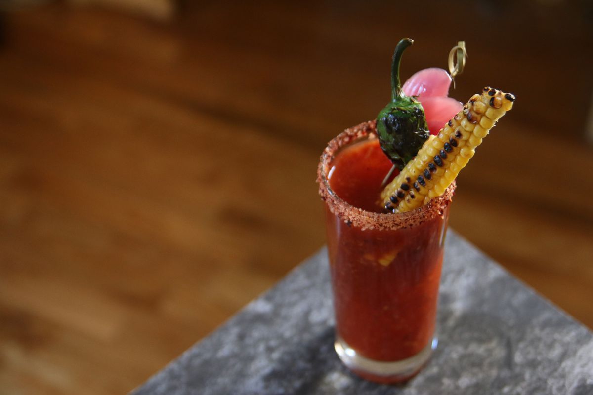 A red-colored cocktail in a tall glass with a chile salt rim and a grilled corn and pepper garnish.