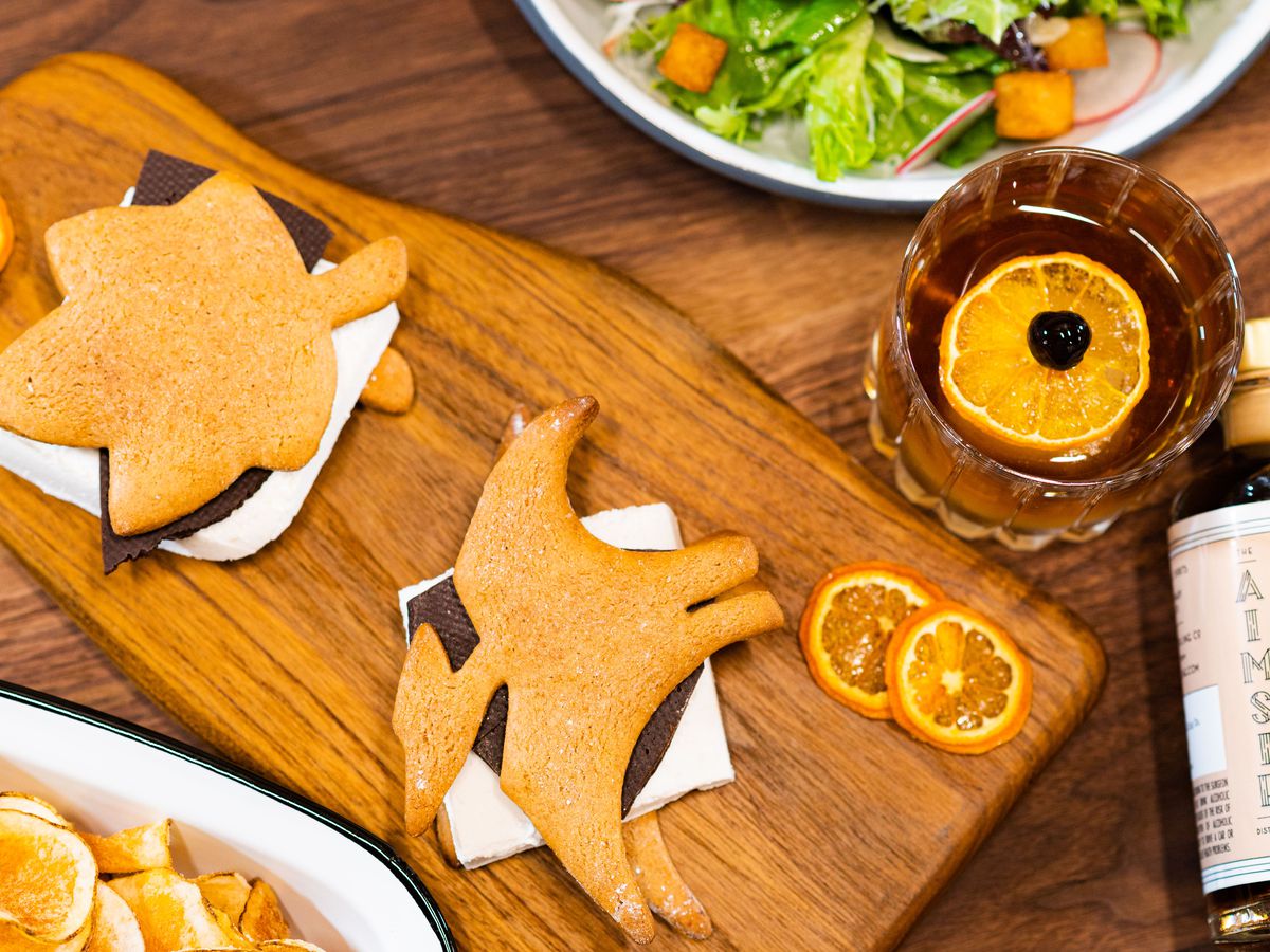 On a wooden table, a wooden paddle holds a pterodactyl-shaped s’more and a leaf-shaped s’more. Next to the paddle is an old fashioned with a large orange wheel. 