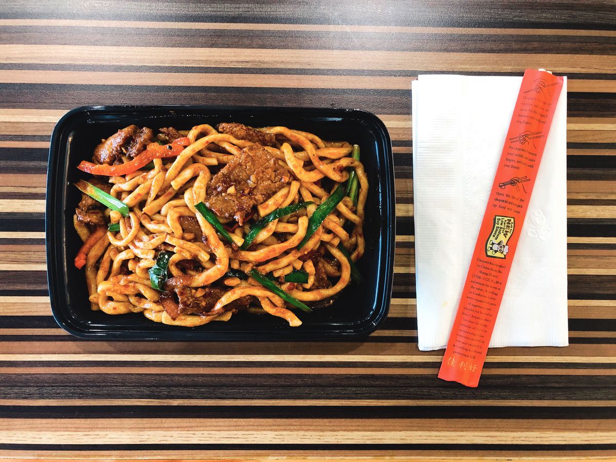 A takeout container of dry-fried noodles with beef at Silk Road Express in Allston, served on a striped wooden table with a pair of chopsticks.