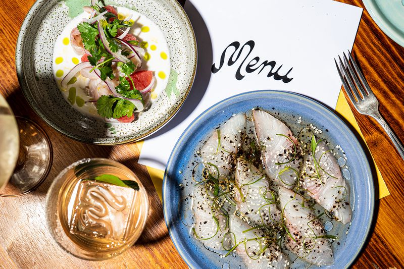 Two modern crudo-style fish preparations sit on a table beside colorful cocktails. 