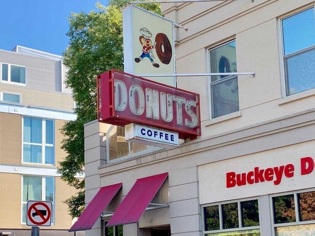 A shop exterior, with a large hanging sign for donuts, another for coffee, lettering indicating the name of the shop on the facade, and an illustrated sign of an anthropomorphized animal boy in a chef’s toque pushing a giant donut.