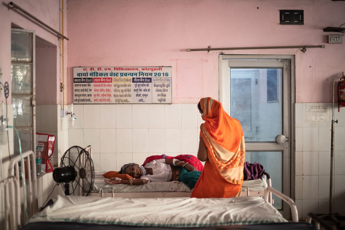 A visitor wearing a sari sits on a bed beside a patient receiving oxygen at a hospital in India.