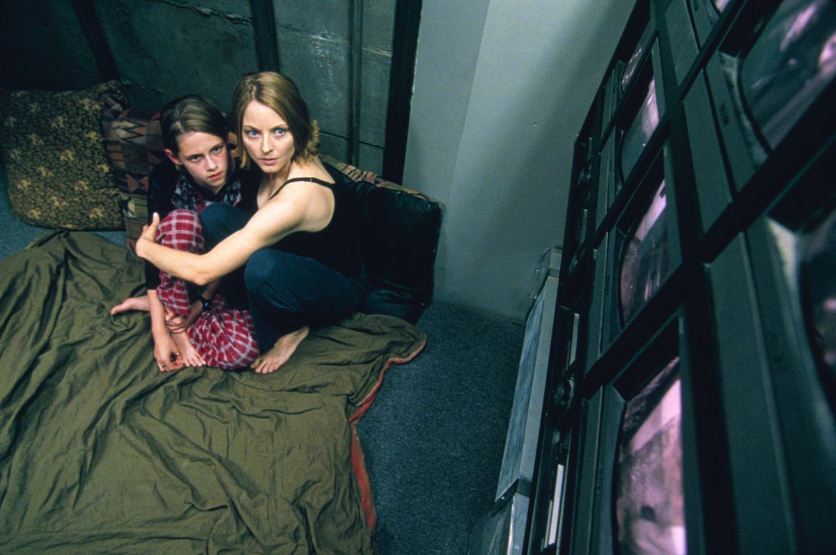 Jodie Foster and Kristen Stewart huddle on a green blanket while watching a wall of screens in Panic Room.
