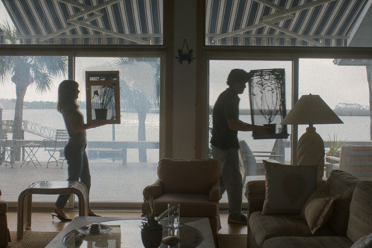 A scene from the movie May December shows a woman and a man, separated by several feet, walking through a home in front of large glass windows, each carrying a plant.
