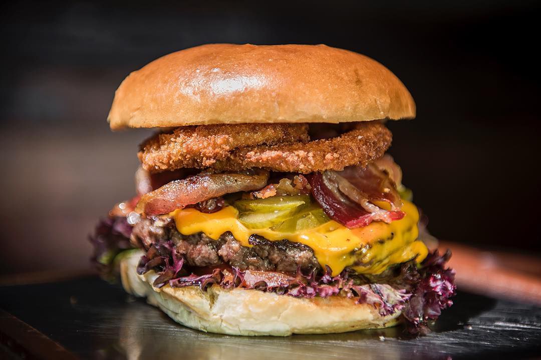 A tall cheeseburger topped with fried onion rings, vegetables, pickles, cheese, and purple lettuce