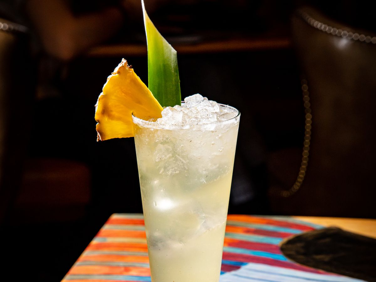 A tall cocktail glass filled with ice and yellow liquid and garnished with a green leaf and pineapple slice.